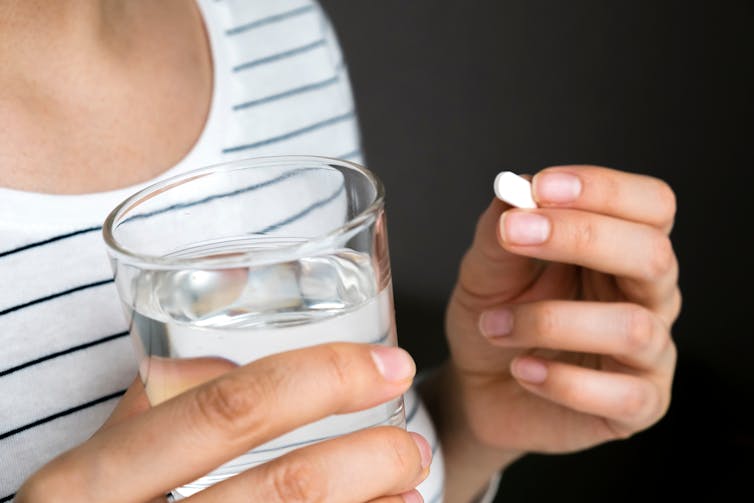 A woman holds a pill in one hand and a glass of water in the other.