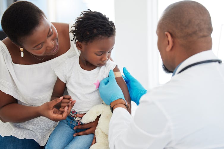 A mother and child with a doctor seen from behind who is putting a bandage on the girl's arm