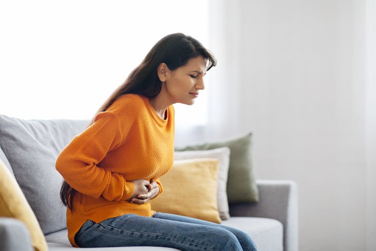 A woman sitting on a couch holds her stomach in pain.