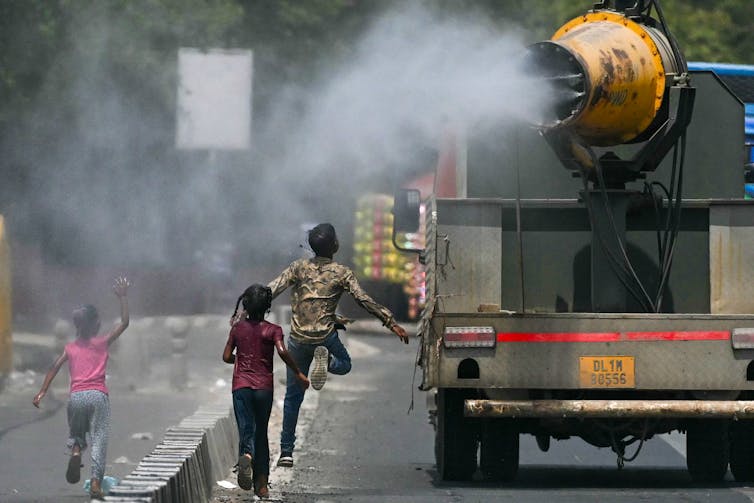 Three children run in the mist being sprayed from a truck. One is leaping for joy into the mist, and a little girl is waving her arms as she runs.