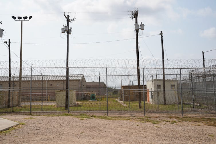 Detainee yard with low buildings behind fences topped with barbed wire and tall light poles