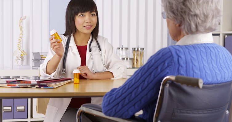 A woman in a lab coat shows medication to an elderly woman, from behind