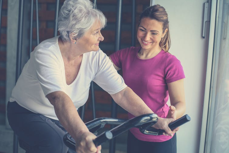 A woman with gray hair on a stationary bike with a fitness trainer in a pink shirt