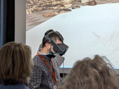 Man with black VR headset on face walks infront of big screen with rocky shore, other people watching in foreground