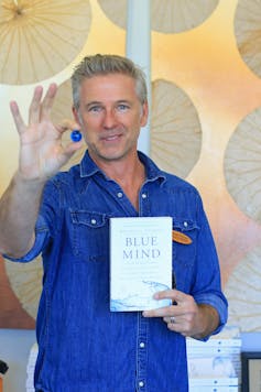 Man in blue shirt holds small blue marble in his right hand and Blue Mind book in his left hand