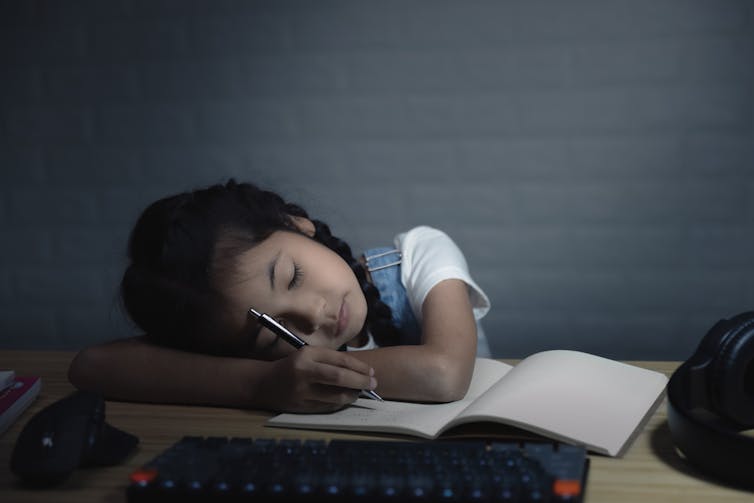 Child resting head in arms against an open notebook, pencil in hand