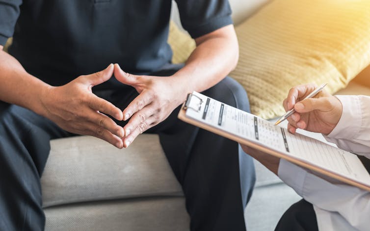 A person is seen sitting, from the shoulders down, beside a medical practitioner who is writing on a clipboard