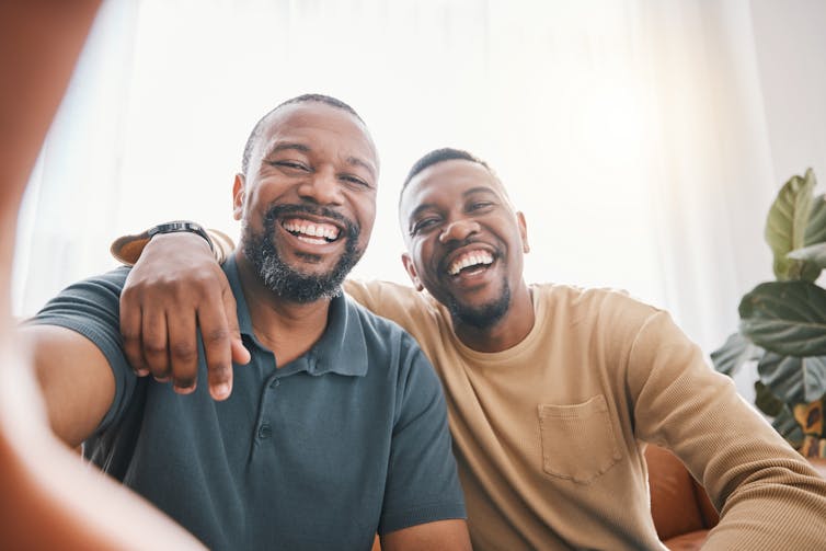 two men sitting next to each other, one has his arm around the other's shoulders, both are smiling