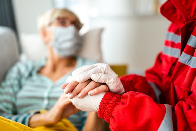 An older woman in an face mask and striped shirt, out of focus, and a paramedic's hands and shoulders.