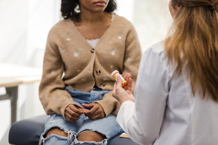 Woman in lab coat holds prescription vial and talks to patient in cardigan and ripped jeans