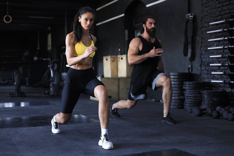 A young man and woman perform lunges in a gym.