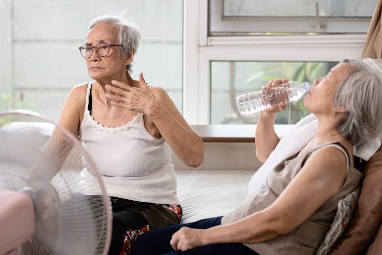 Two older women in front of an electric fan; one is fanning herself with her hand and the other is drinking water