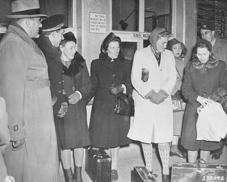 Four women in winter coats stand with suitcases, with a few men standing around them.