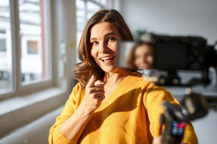 A young woman in a bright yellow sweater smiles and points her finger at a videocamera she is holding.