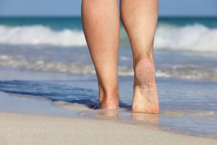 woman bare legs and feed in shallow sea water walking along sandy beach, white waves in background