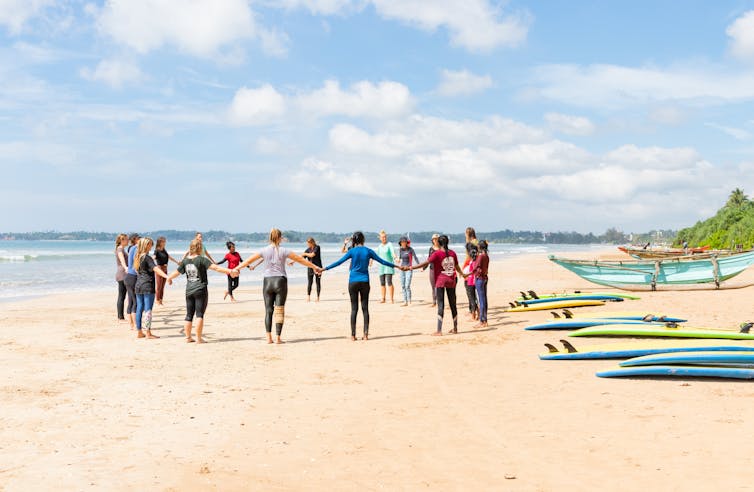 tropical beach, group of women holding hands on sand, surfboards on beach, calm ocean in background, blue sky