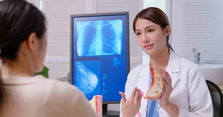 A female doctor holds a breast model up, explaining it to her patient, with a mammogram image in the background.