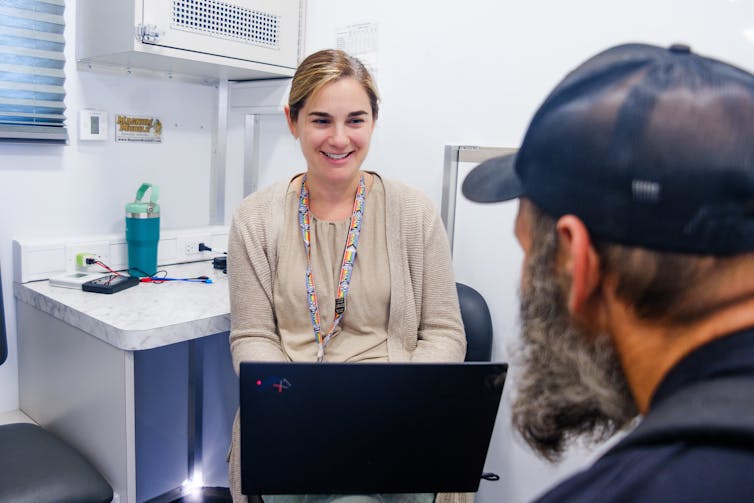 A smiling woman with laptop meets sits across from man with black baseball cap and beard