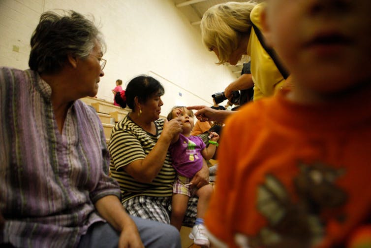 Person holding child, sitting by two other people; in the foreground, a child approaches the camera