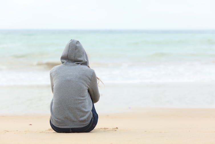 A person with a hoodie on sitting on the beach, staring out to sea.