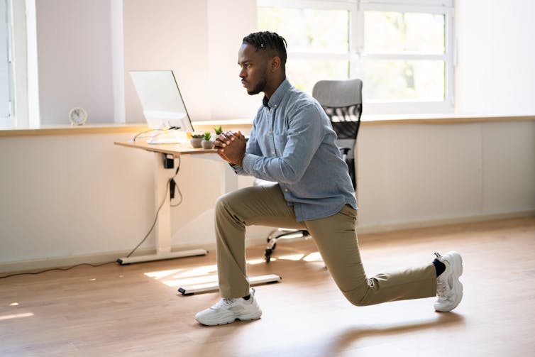 A man wearing business casual attire performs a lunge in his office.