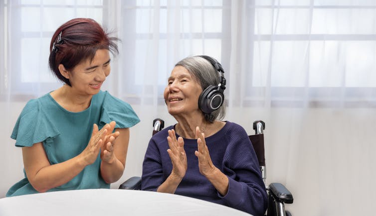 A woman with dementia listens to music with headphones, while her nurse or caregiver sits beside her.