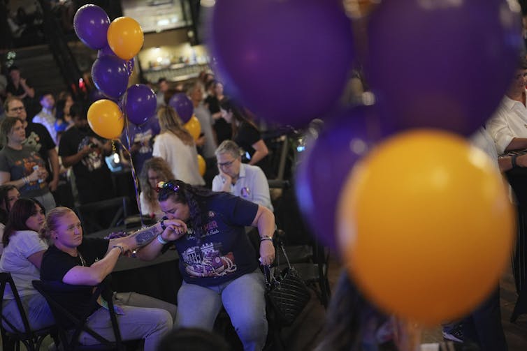 A woman kisses the hand of another woman who looks crestfallen. Purple and orange balloons are in the foreground.