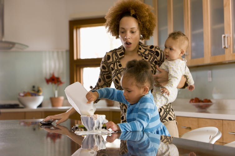 Young child spills milk all over the counter as mother holding an infant looks on in disappointment and distress.