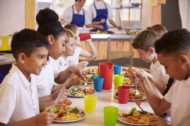 primary schoolchildren in white T shirts sitting at table eating healthy food in school canteen, cooks in background