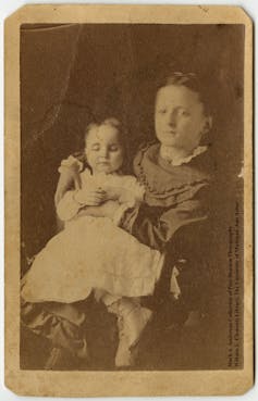 A young girl, perhaps eight years old, dressed in a dark dress, holds a younger girl, possibly three years old and dressed in white, on her lap