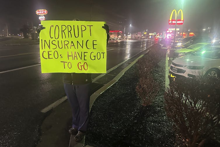 person holds up yellow sign with words 'corrupt insurance ceos have got to go' while standing on sidewalk near street at night with mcdonald's arches in background