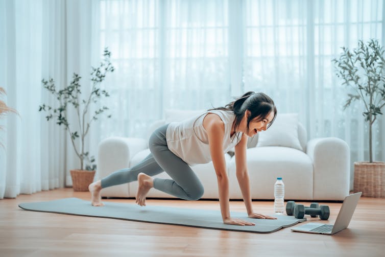 A woman performs a mountain climber exercise on her yoga mat while following a workout on her laptop.