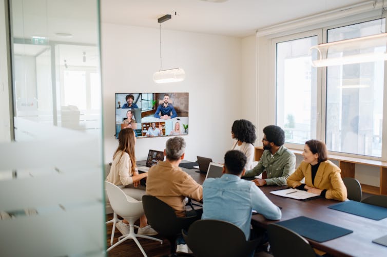 A group of people sitting at a conference table look at a screen mounted on a wall that shows a video call with several people