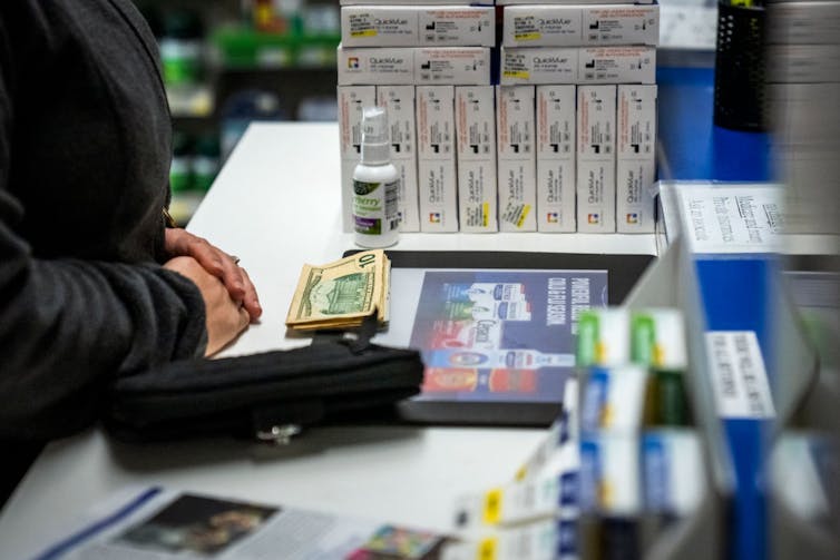 Customer at counter of pharmacy, a stack of bills and drug packages flanking