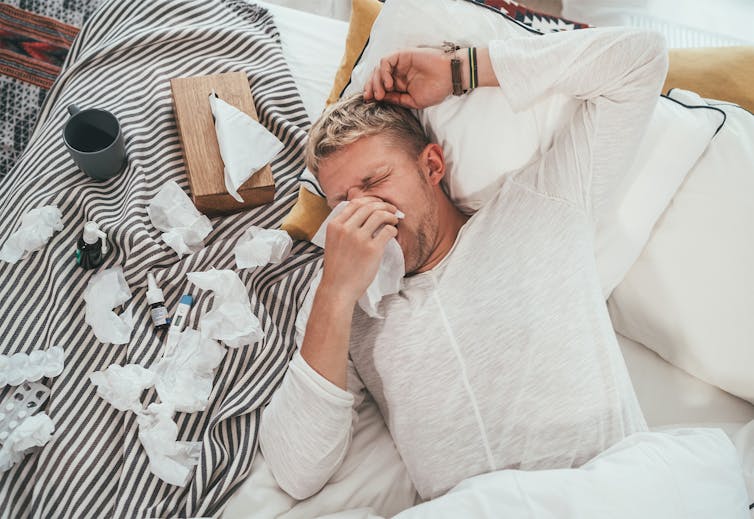 A sick man lays in bed while blowing his nose.