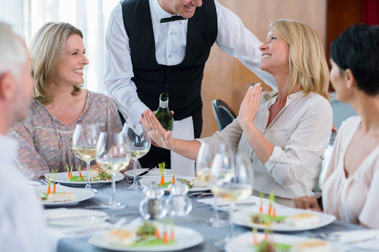 A group of people dine at a restaurant. One woman refuses another glass of wine from a waiter.