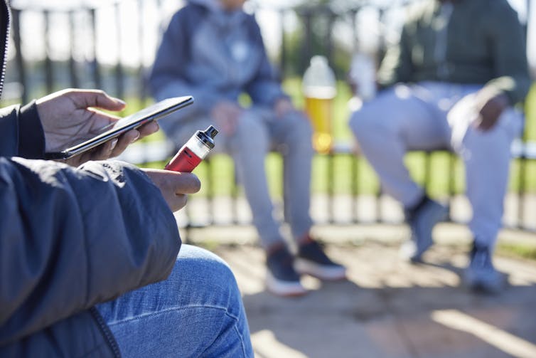 Close-up of teenagers sitting on benches, one holding a vape cartridge and cell phone in foreground.