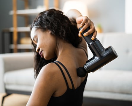 A woman using a massage gun on her back in what looks like her living room