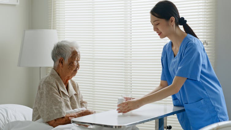 A young woman in blue scrubs with an older woman sitting in bed