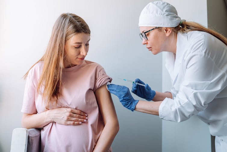 Pregnant woman holding her abdomen, getting vaccine injection in her arm from doctor syringe.