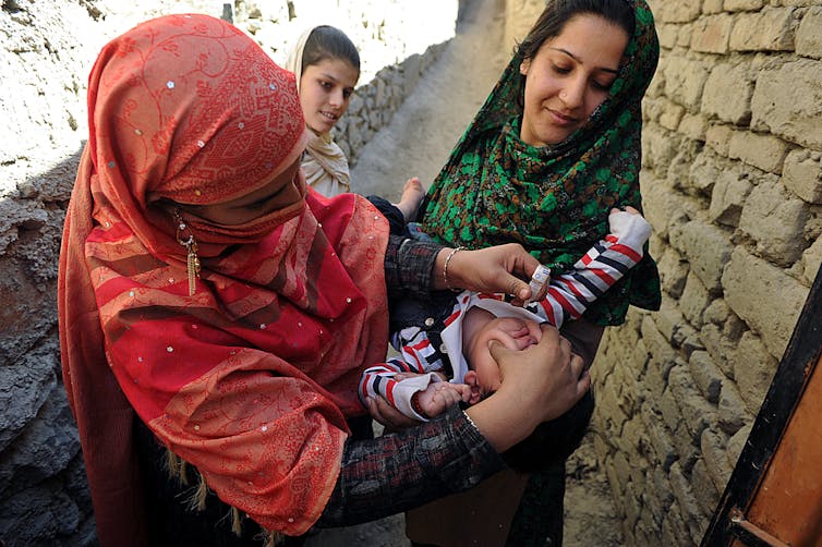Woman in traditional Afghan dress tries to squeeze medical drops into mouth of a baby held in another woman's arms