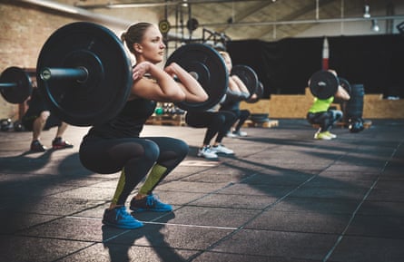 Training together and staying focusedFit young woman lifting barbells looking focused, working out in a gym with other people