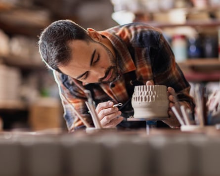 Young man potter making pattern on clay mug with special tool in pottery workshop