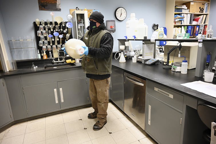 A man wearing a mask and gloves shakes a large jug in a room with lab equipment.