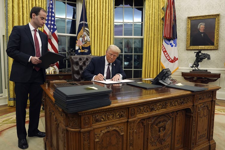 A man signing a document behind a large desk while another man watches