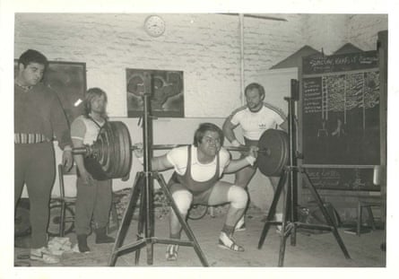 A black and white photograph of Arthur White squatting while holding a barbell with three other men looking on