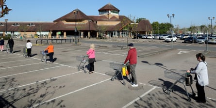 People queue up in a supermarket car park, standing well apart from each other