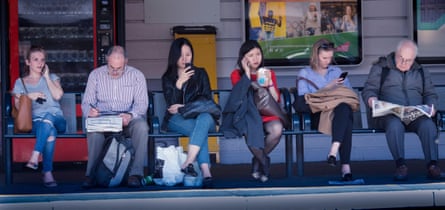 People sitting at a train station, most staring at their phone or talking on it, a couple reading the paper