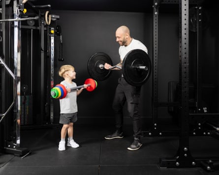 Rich Hawkins holding a large black barbell, looking at his son Saxon holding a small colourful barbell
