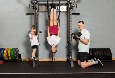 Ottilie hangs upside down on a training machine holding hoops, with Atticus standing on one side holding dumbbells and Alex kneeling on the other holding large dumbbells